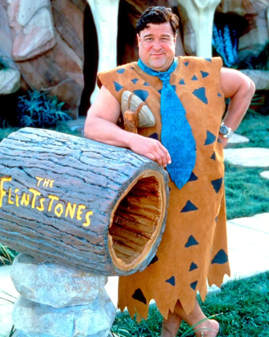 John Goodman stands in costume as Fred Flintstone, leaning against a stone mailbox on the set of The Flintstones.