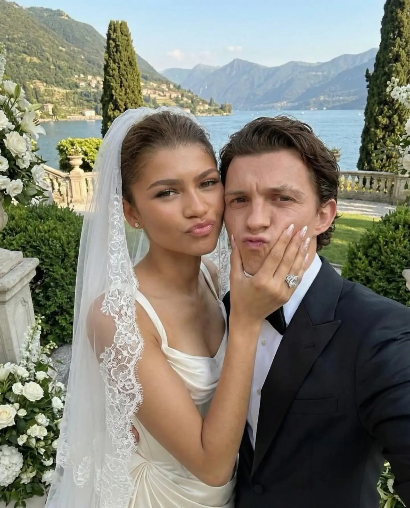 AI-generated selfie of Zendaya in a lace bridal veil and Tom Holland in a tuxedo making playful faces by a scenic lake.