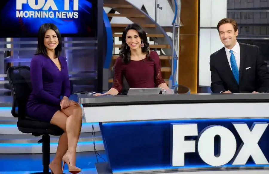 Tracee Carrasco sits on a news set in a purple dress alongside two colleagues behind a FOX news desk.