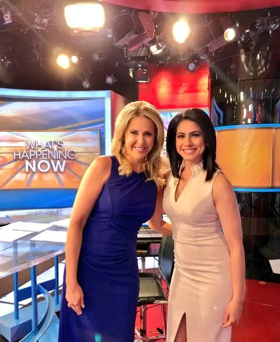 Cheryl Casone in a blue dress and Tracee Carrasco in a sleeveless white dress smile together on the "What