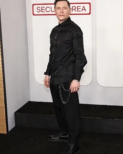 Burn Gorman poses on a black carpet wearing an all-black outfit featuring a patterned button-down shirt and trousers with a silver wallet chain.