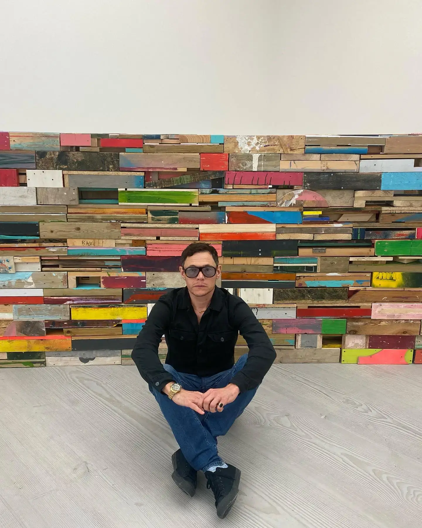 Burn Gorman sits cross-legged on a light wood floor in front of a colorful, abstract wall made of reclaimed wood planks.