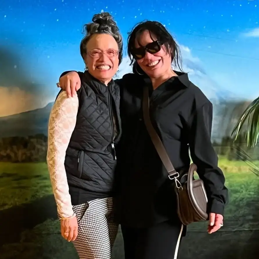 Rachel House and Rena Owen smile warmly with their arms around each other in front of a scenic mountain backdrop.