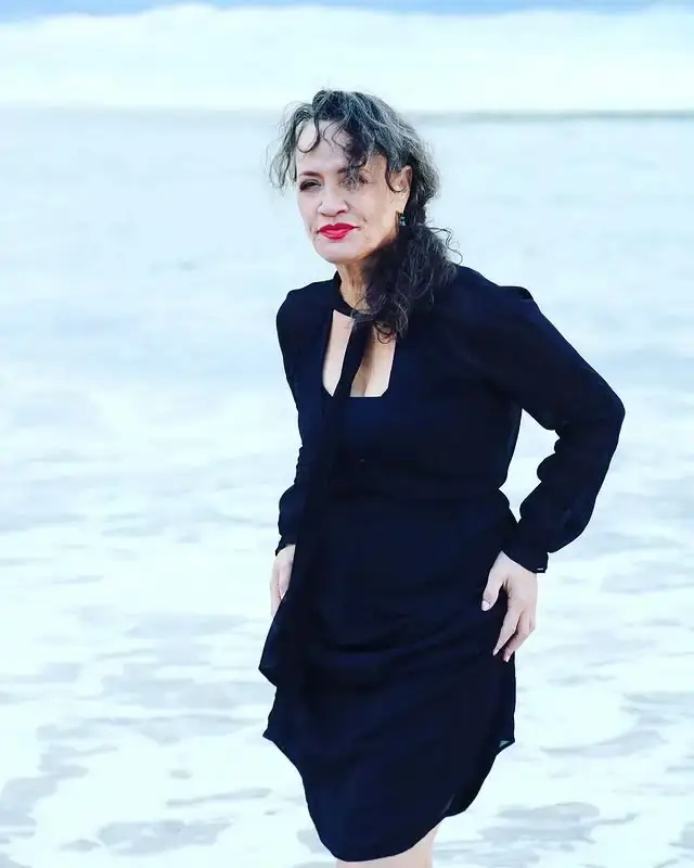 Rena Owen posing on a beach in a black dress with the ocean waves blurred in the background.