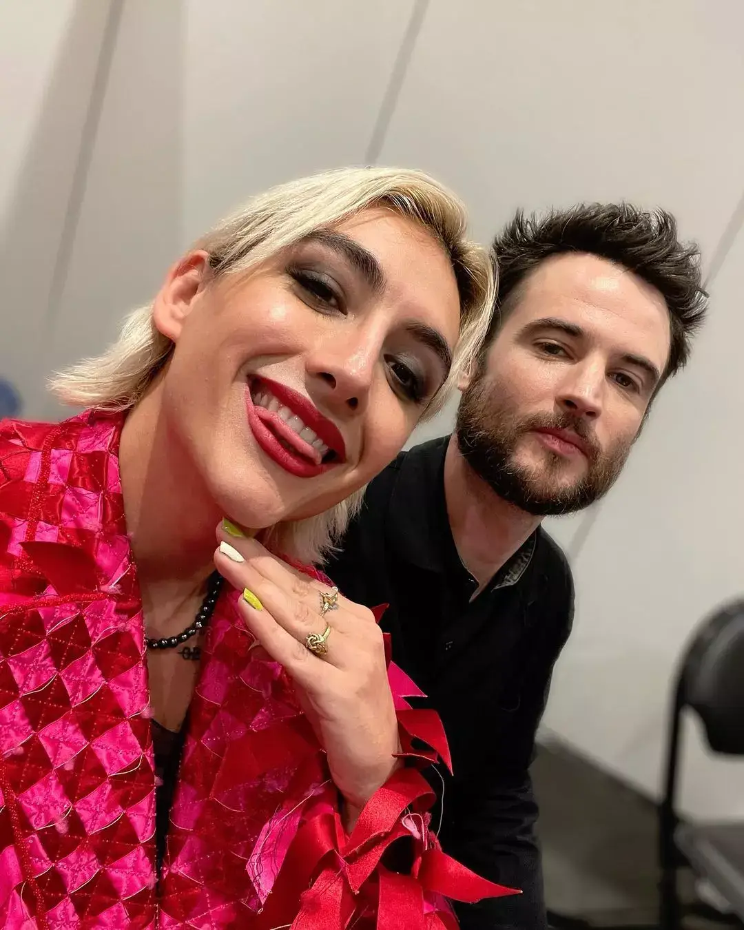 Mason Alexander Park, in a textured red jacket, takes a close-up selfie with a bearded Tom Sturridge.