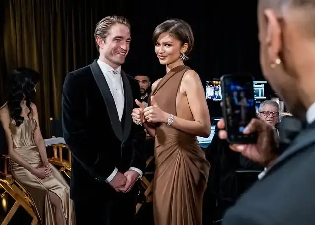 Zendaya, in a chocolate-brown one-shoulder gown, and Robert Pattinson, in a black velvet tuxedo, stand together and smile at the 2026 Oscars.