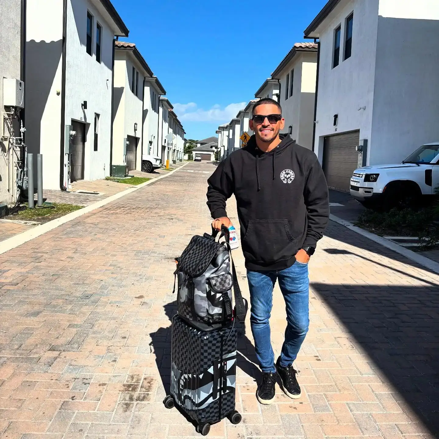 Boxing champion Jorge Linares stands on a sunlit residential street wearing a black hoodie and sunglasses, posing next to his designer luggage.