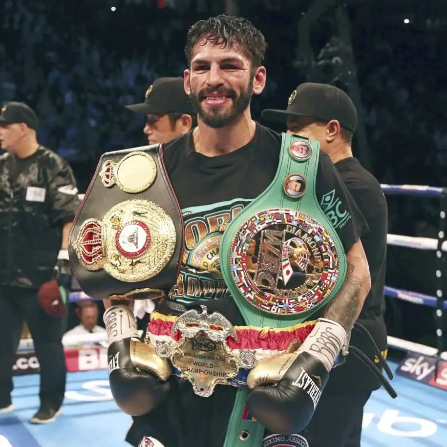 A smiling Jorge Linares stands in a boxing ring, draped in multiple championship belts including the WBC Diamond and WBA world titles.
