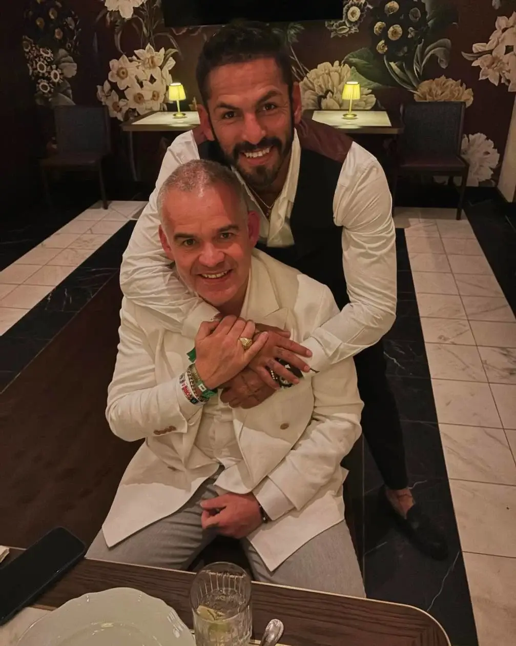 Jorge Linares stands behind Gilberto Jesús Mendoza, hugging him around the shoulders as they both smile warmly at a dinner table in a restaurant.