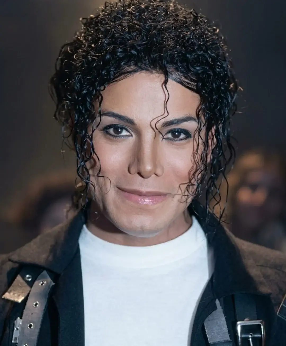 Jaafar Jackson, dressed as Michael Jackson in a white V-neck shirt and black trousers, performs an iconic dance move on stage during filming.