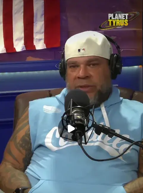 Tyrus sits in a leather chair wearing a white baseball cap and a light blue sleeveless hoodie while speaking into a professional studio microphone.