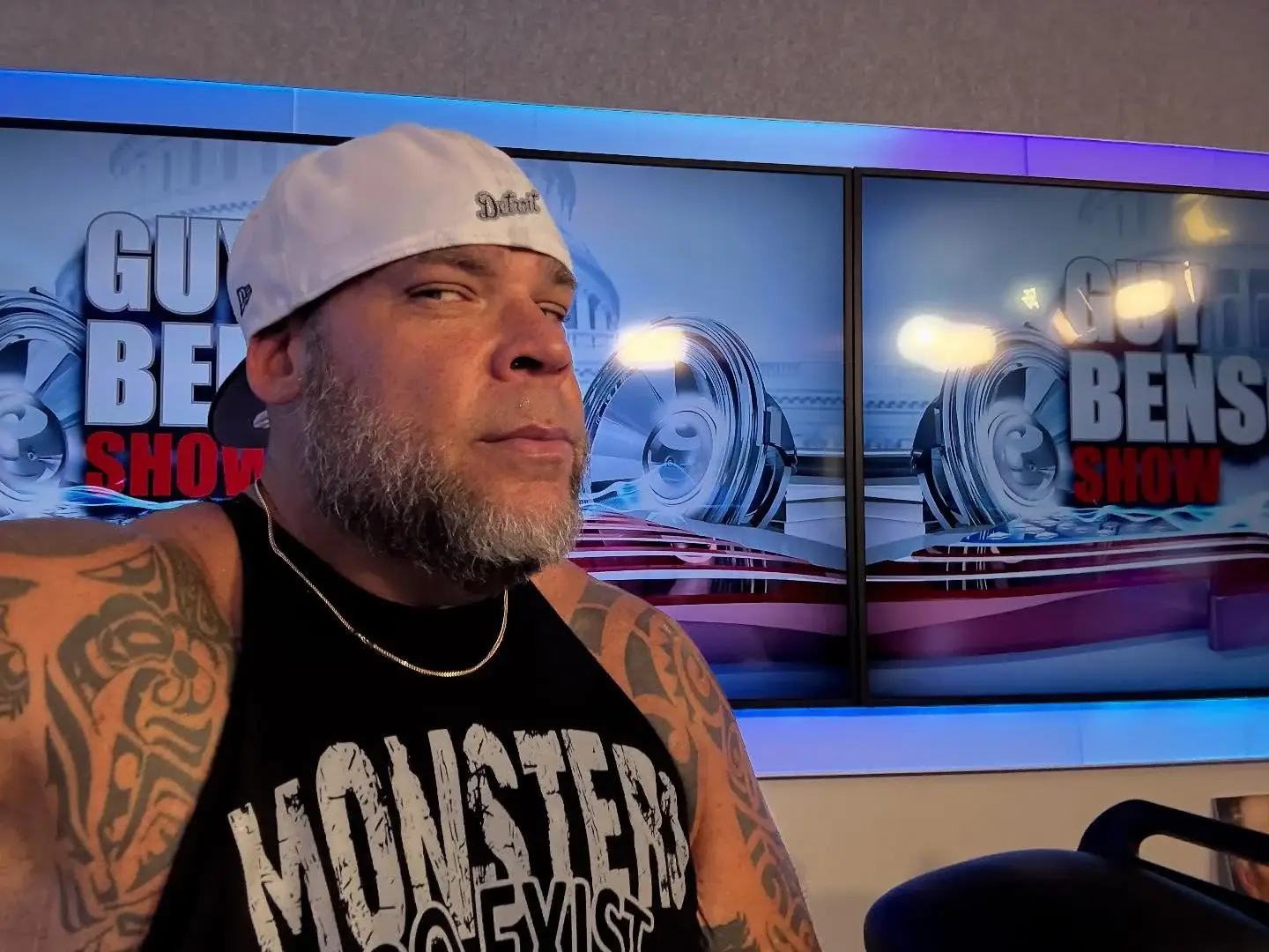 A selfie of Tyrus wearing a white Detroit baseball cap and a black "Monster" tank top in front of The Guy Benson Show studio monitors.