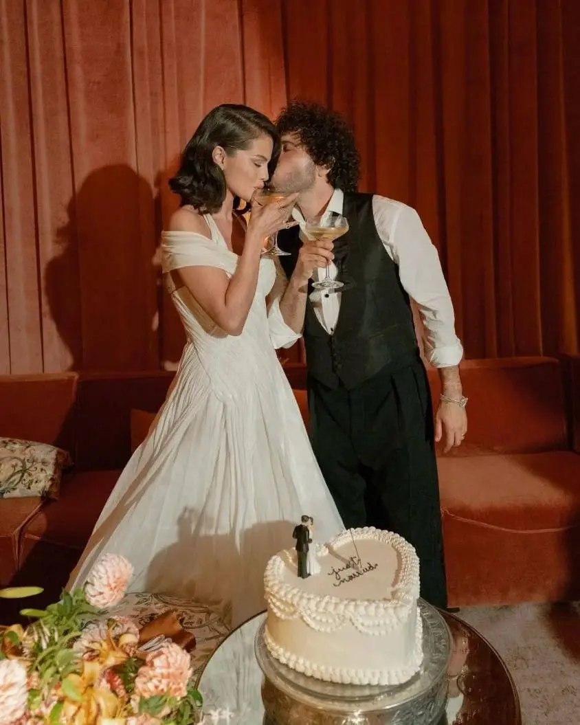 Selena Gomez in a white gown and Benny Blanco in a tuxedo toast with champagne glasses behind a heart-shaped "Just Married" cake.