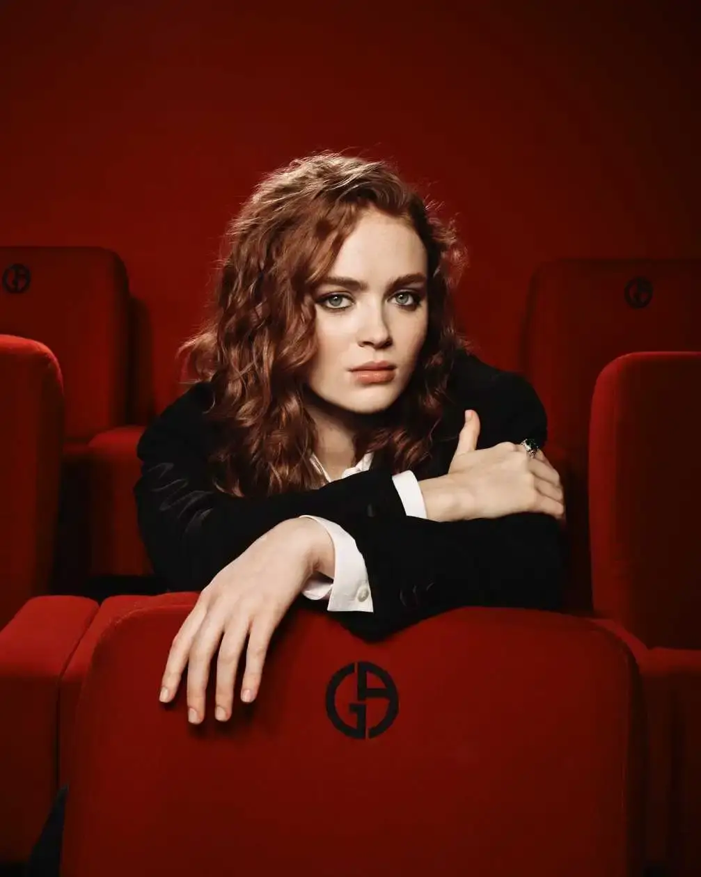 Actress Sadie Sink posing in a black blazer while leaning over red cinema seats with "GA" logos for Armani Beauty.
