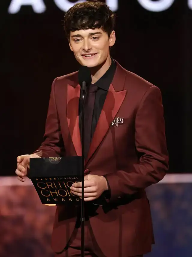Actor Noah Schnapp smiling on stage in a burgundy suit while holding a trophy at the Critics Choice Awards.
