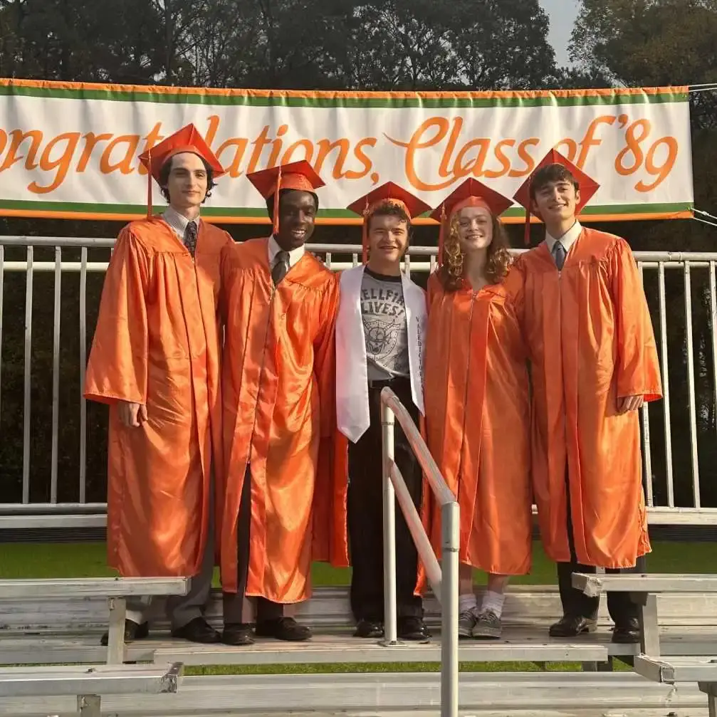 The Stranger Things cast members Finn Wolfhard, Caleb McLaughlin, Gaten Matarazzo, Sadie Sink, and Noah Schnapp posing in orange graduation robes.