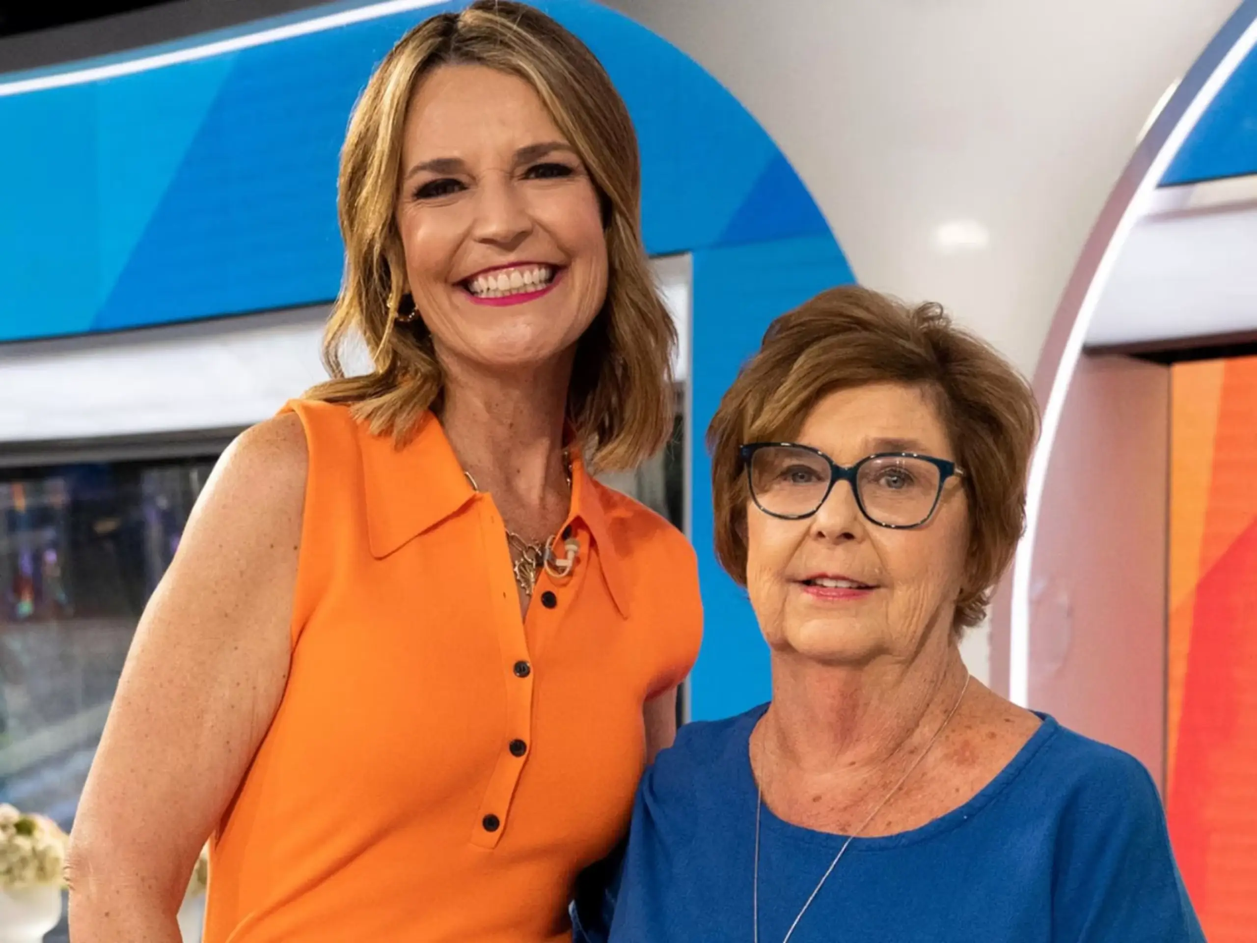 Savannah Guthrie in an orange sleeveless shirt smiling next to her mother, Nancy Guthrie, who is wearing a blue top and glasses.