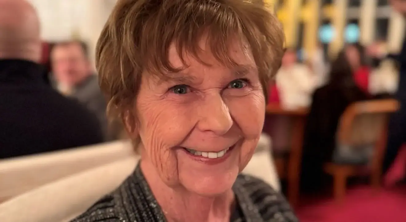 A close-up portrait of Nancy Guthrie smiling warmly while sitting in a dimly lit indoor setting.