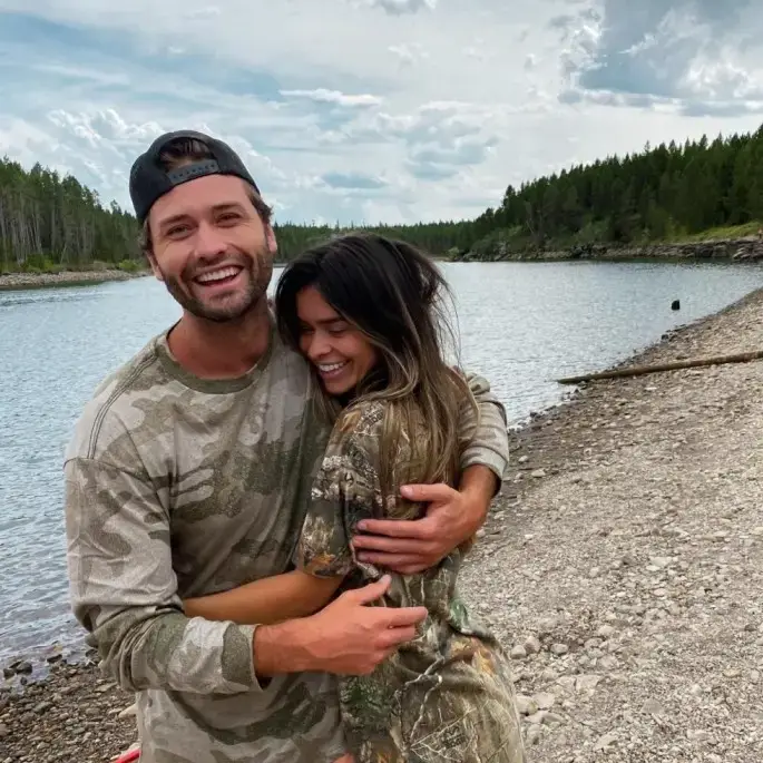 Dakota Mortensen and Taylor Frankie Paul hugging and laughing while wearing matching camouflage shirts on a rocky lakeshore.