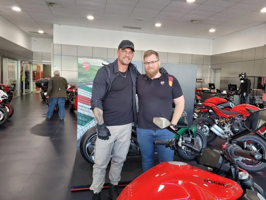 Alan Ritchson poses with a fan in a Ducati motorcycle showroom surrounded by red sportbikes.