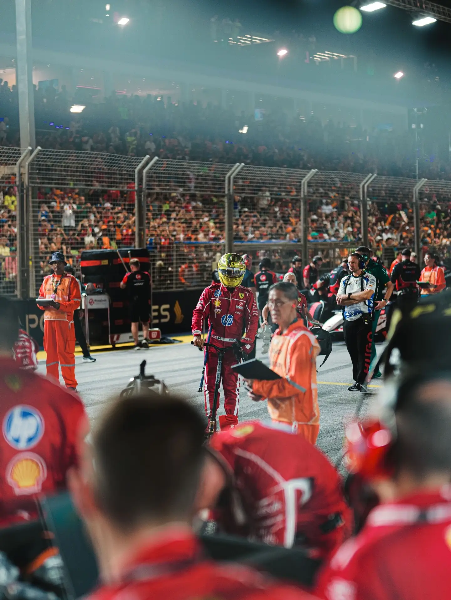 Lewis Hamilton stands on a crowded Formula 1 grid at night wearing a red Ferrari racing suit and a yellow helmet.