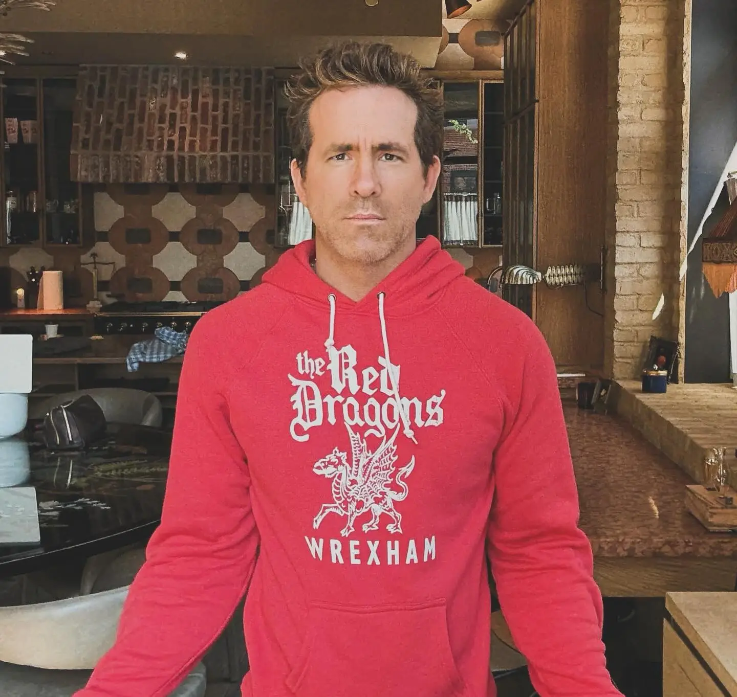 Ryan Reynolds wearing a red Wrexham AFC "The Red Dragons" hoodie in a kitchen setting.