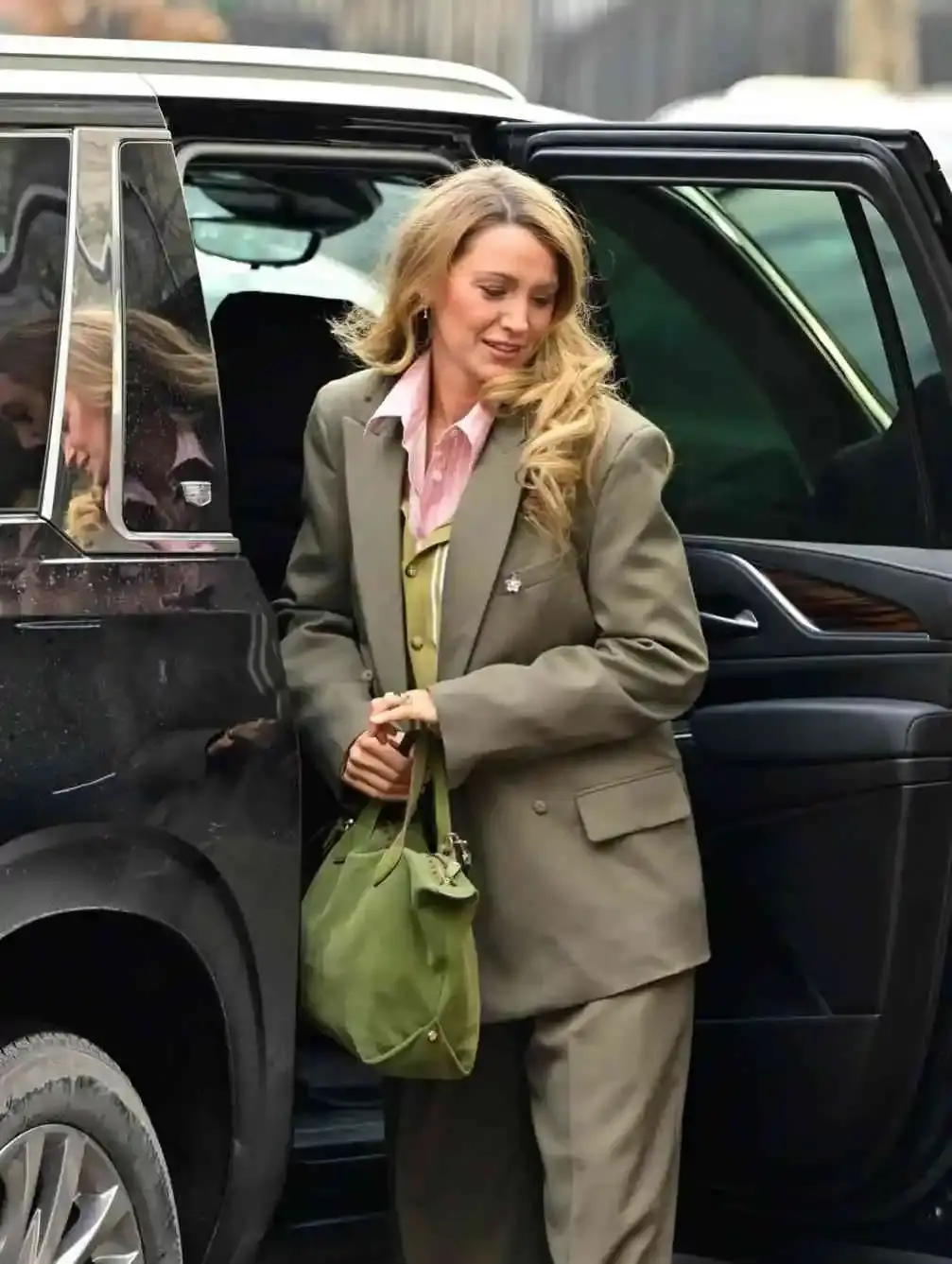 Blake Lively exiting a black vehicle while wearing an oversized olive green suit over a pink button-down shirt.