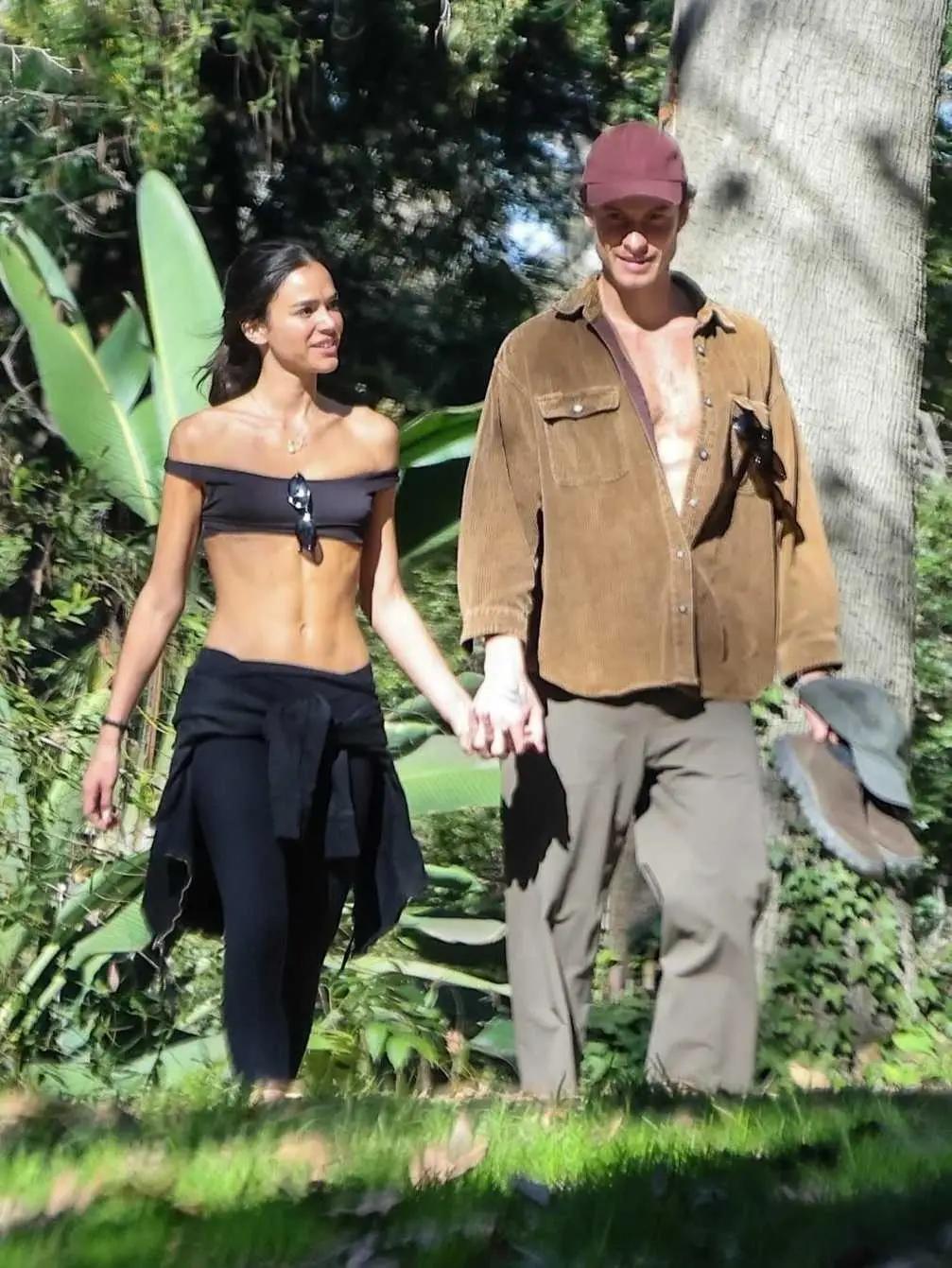 Shawn Mendes and Bruna Marquezine hold hands while walking together through a sunlit, wooded park.