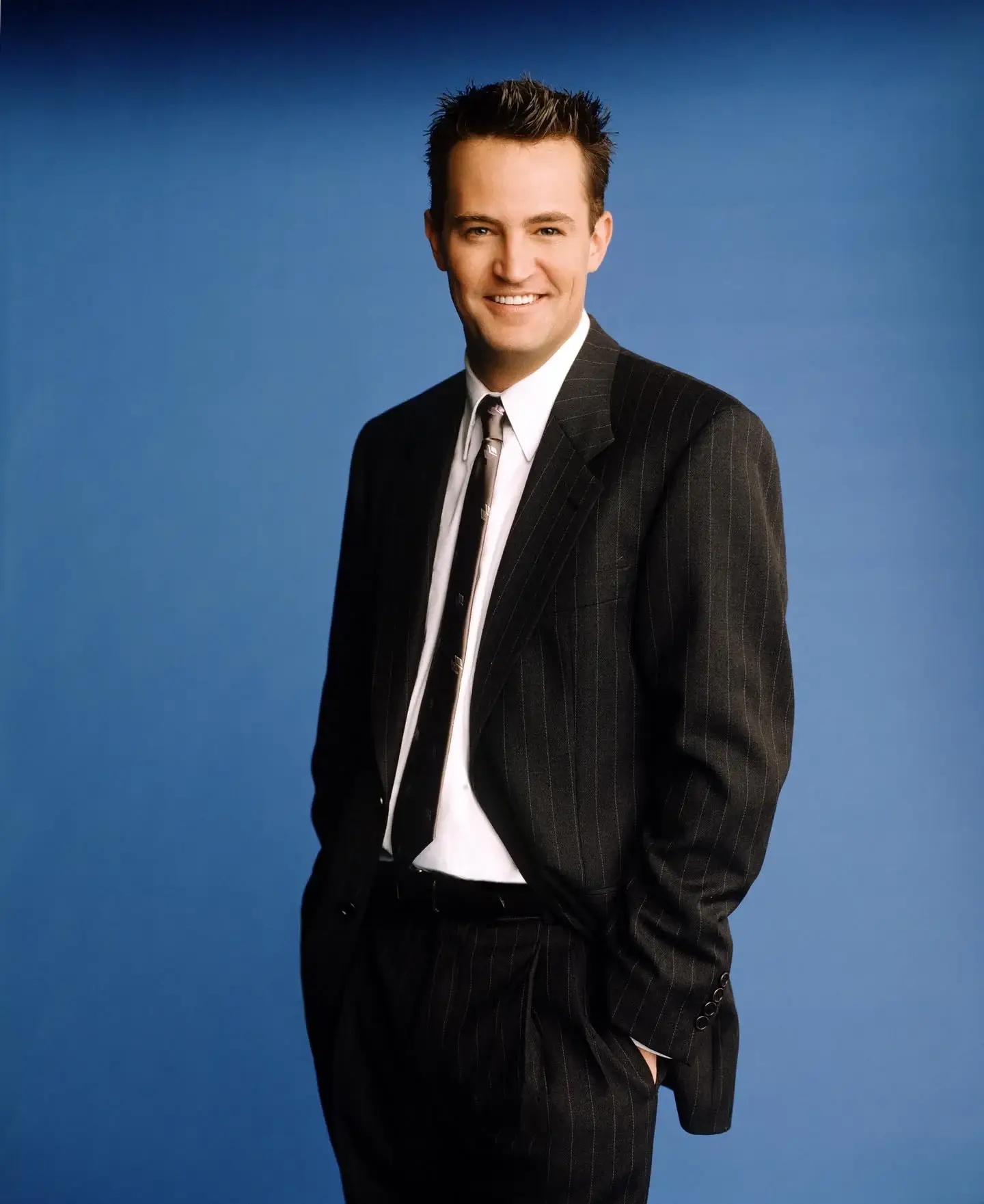 Matthew Perry stands smiling on a dimly lit street, looking relaxed in a dark tailored suit.