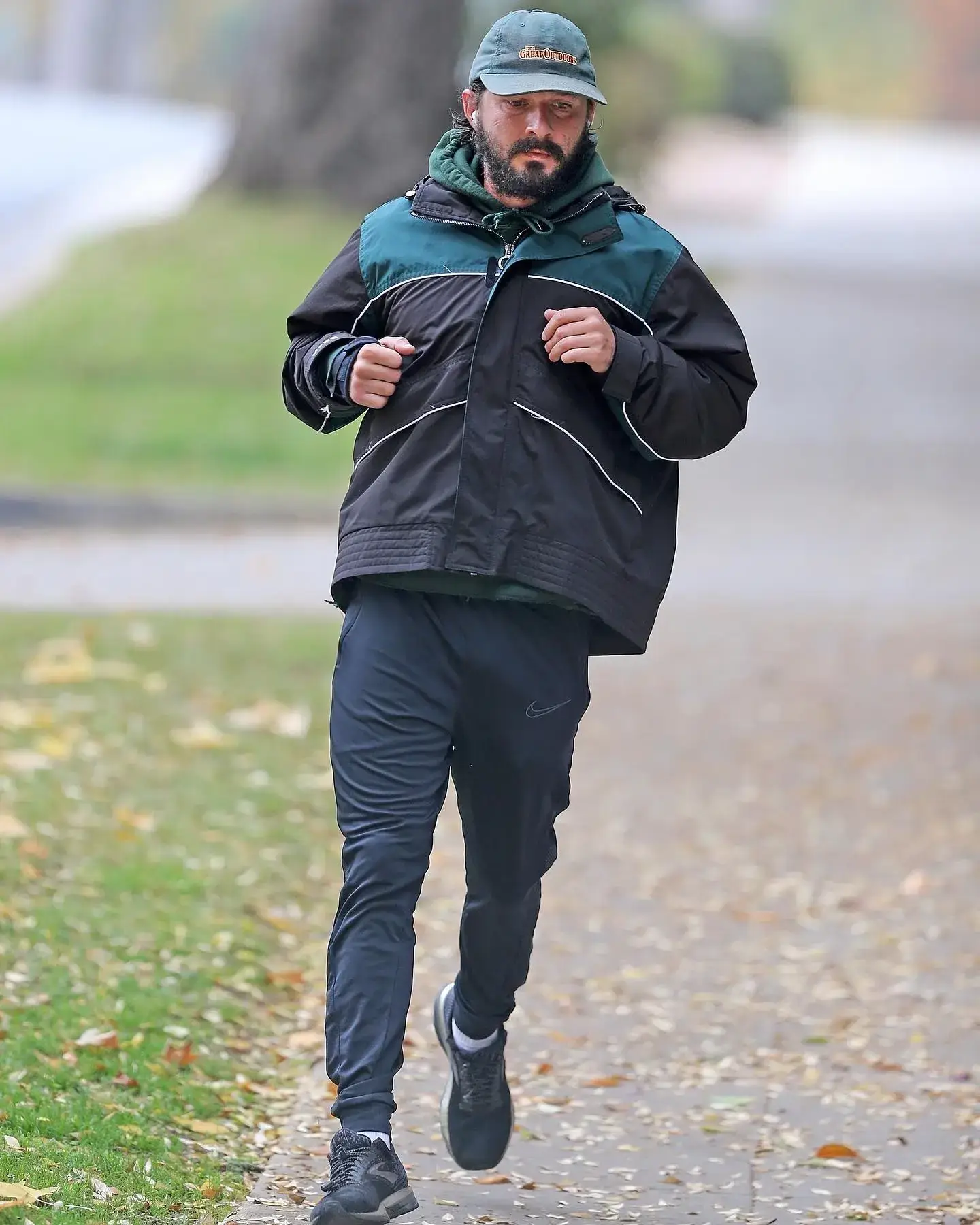 Shia LaBeouf jogging outdoors wearing a green cap, a black and teal jacket, and athletic pants.