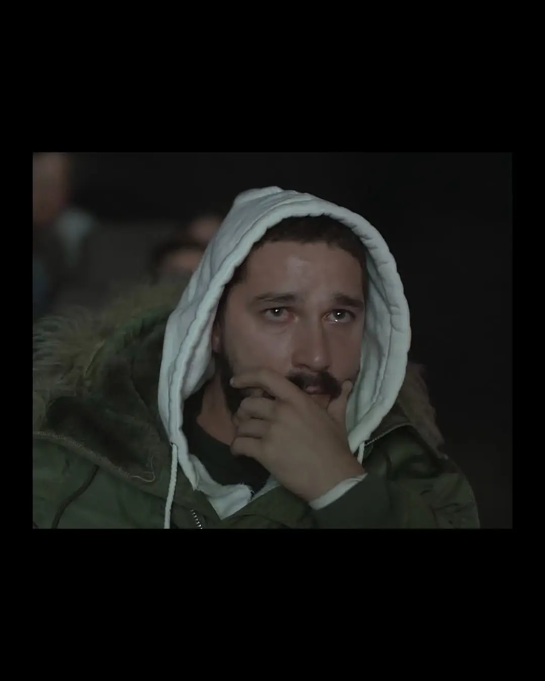 Shia LaBeouf wearing a white hoodie and a green parka while looking contemplative and emotional.
