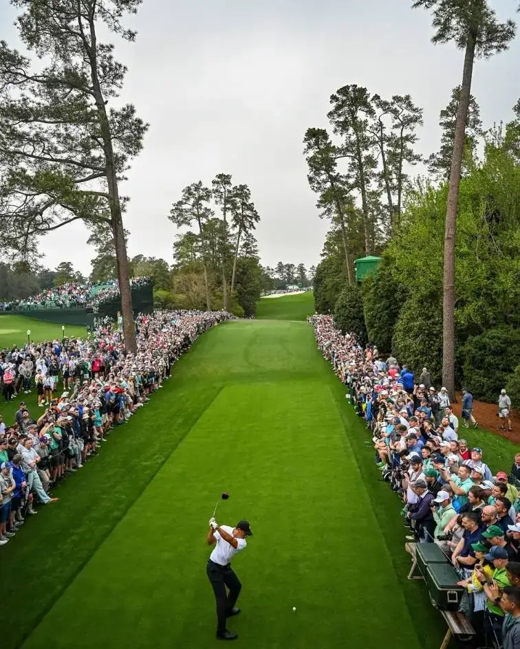 Tiger Woods mid-swing on a narrow, tree-lined fairway surrounded by a massive gallery of spectators.
