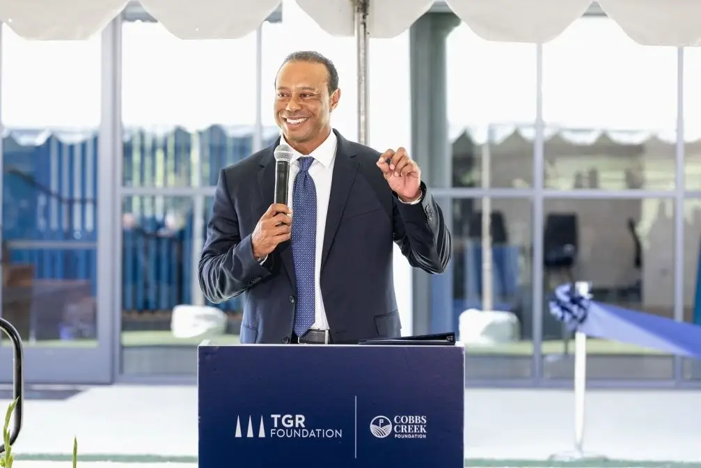 Tiger Woods smiling and speaking into a microphone at a TGR Foundation podium.