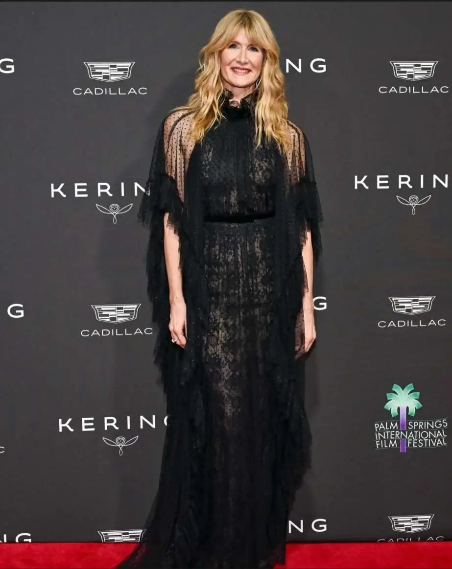 Laura Dern smiling on a red carpet wearing a high-neck, ruffled black lace gown with sheer polka-dot sleeves.