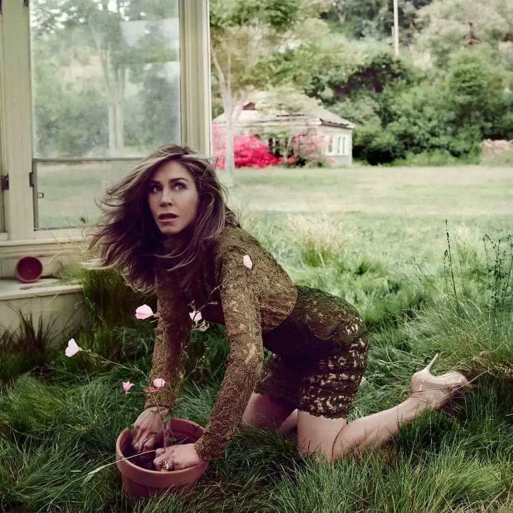 Jennifer Aniston poses in a green lace dress while kneeling in the grass next to a potted plant.