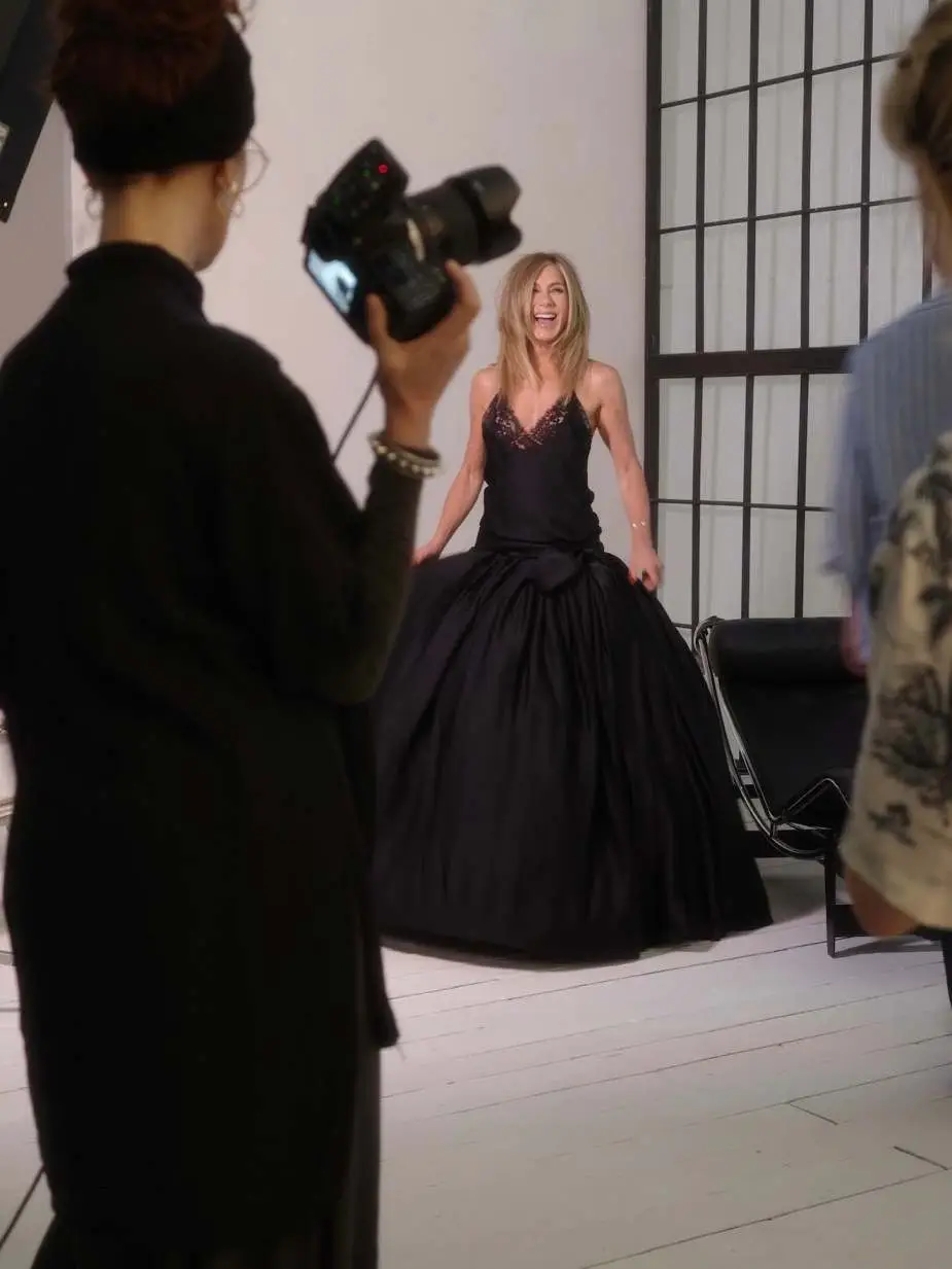 Jennifer Aniston laughs during a photoshoot while wearing a voluminous black gown with a lace bodice.