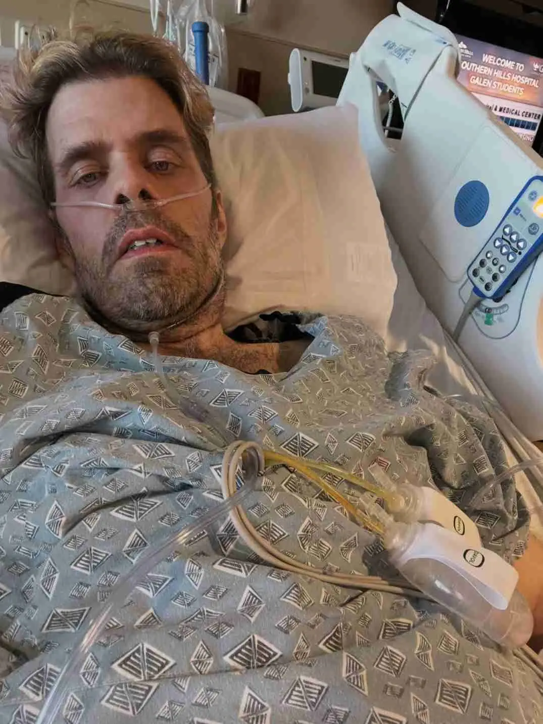 Perez Hilton lies in a hospital bed wearing a patterned gown and nasal cannula for oxygen support.