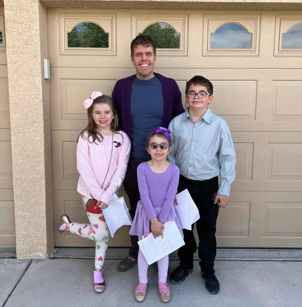 Perez Hilton stands smiling in front of a garage door with his three children, who are dressed up and holding papers.
