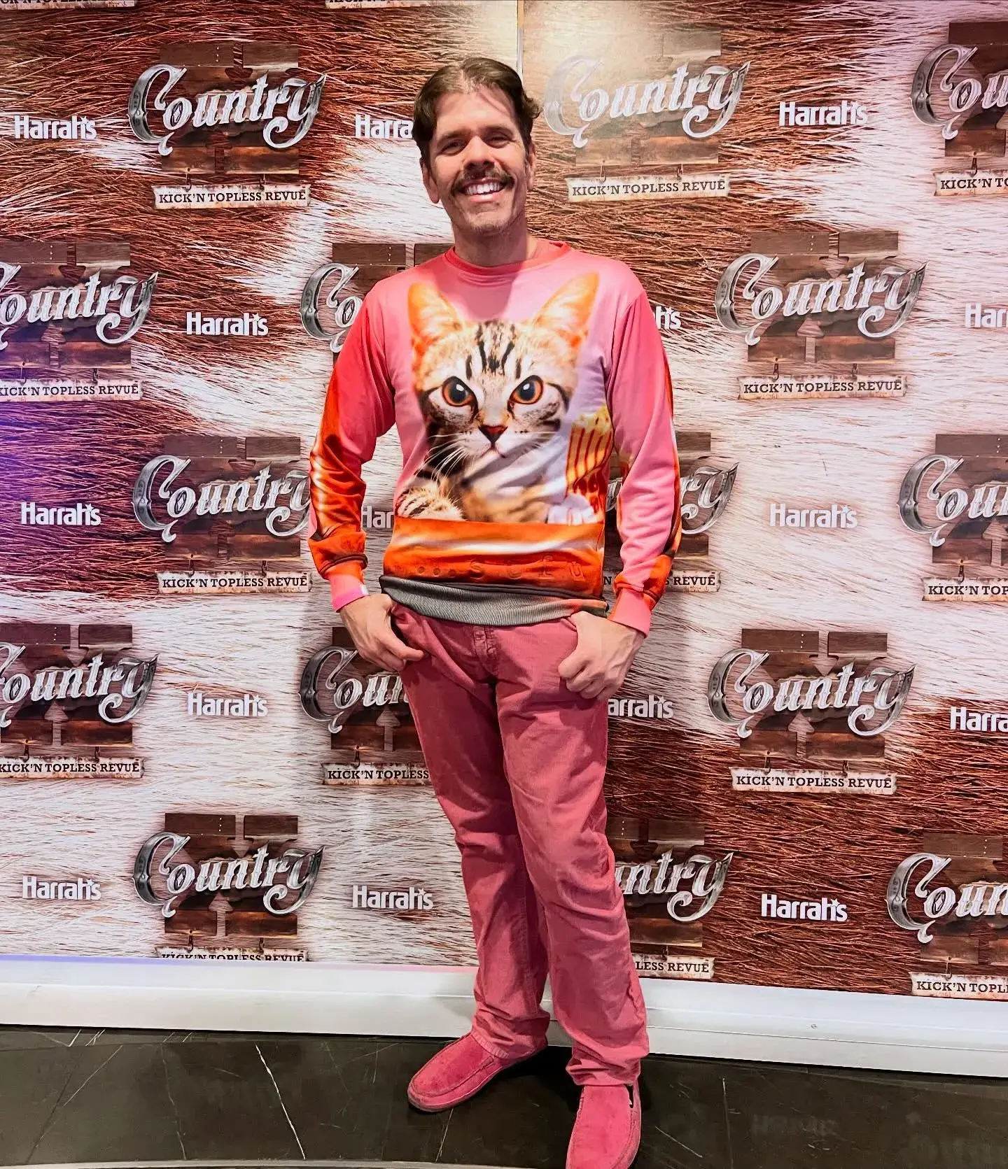 Perez Hilton poses on a red carpet wearing a pink sweatshirt with a large cat graphic and matching pink trousers.