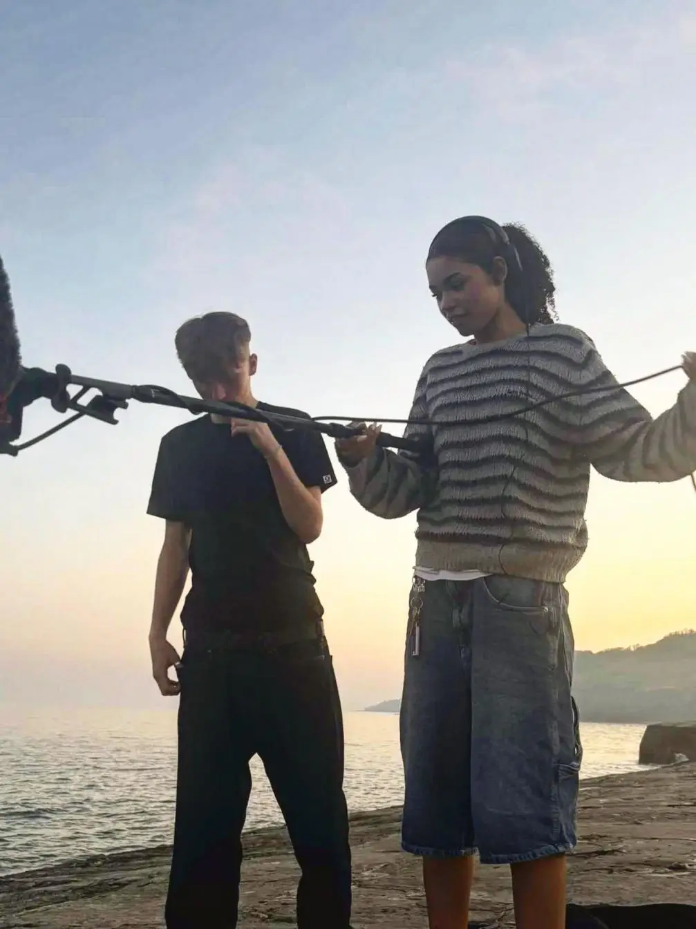 Ida Brooke wears a striped sweater and denim shorts while handling audio equipment on a coastal film set.