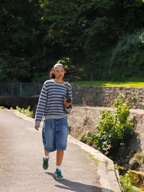 Ida Brooke walks along a sunny path wearing a striped sweater, denim shorts, and green sneakers.