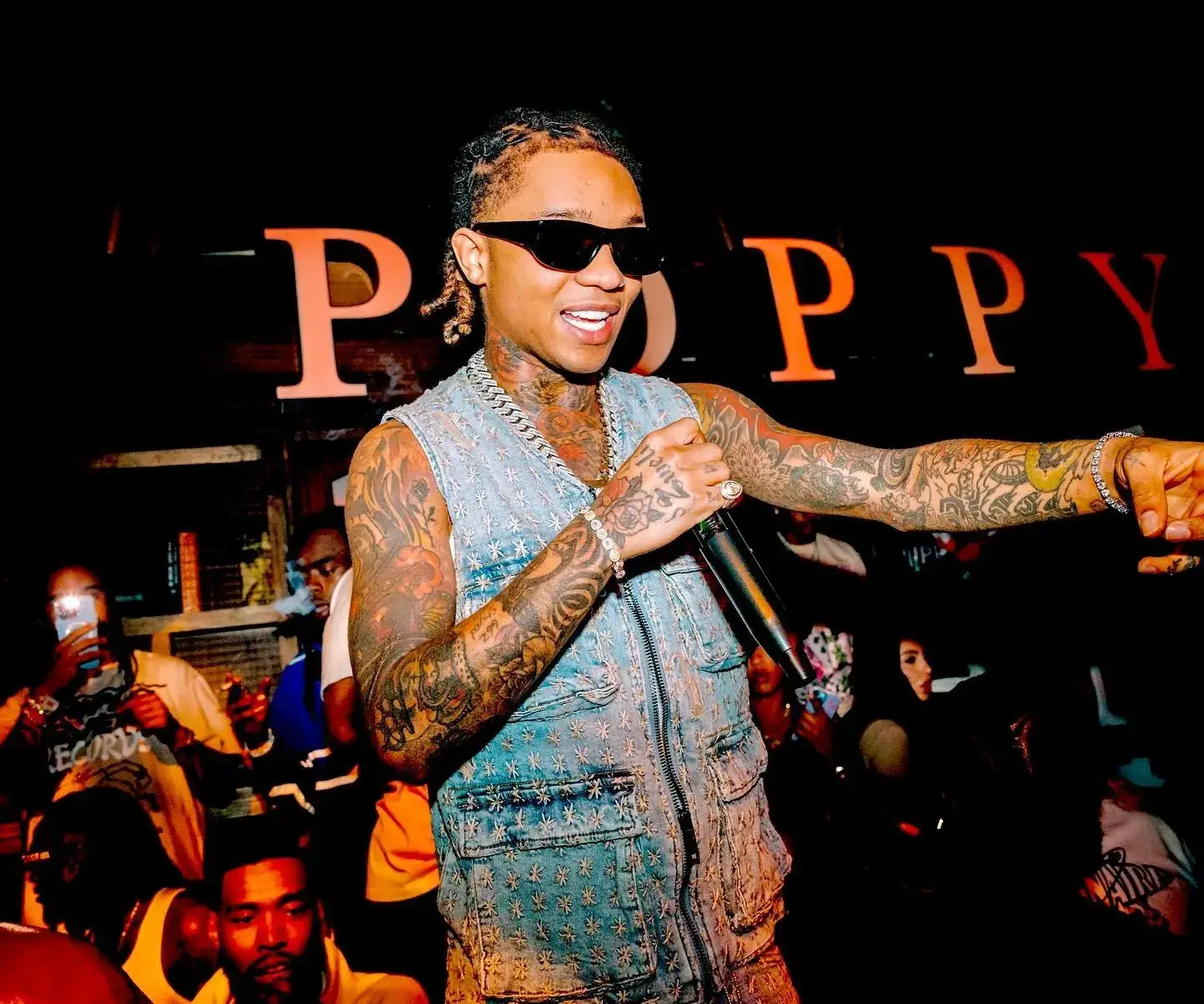 Swae Lee performs with a microphone in a denim vest and sunglasses during a vibrant live concert event.