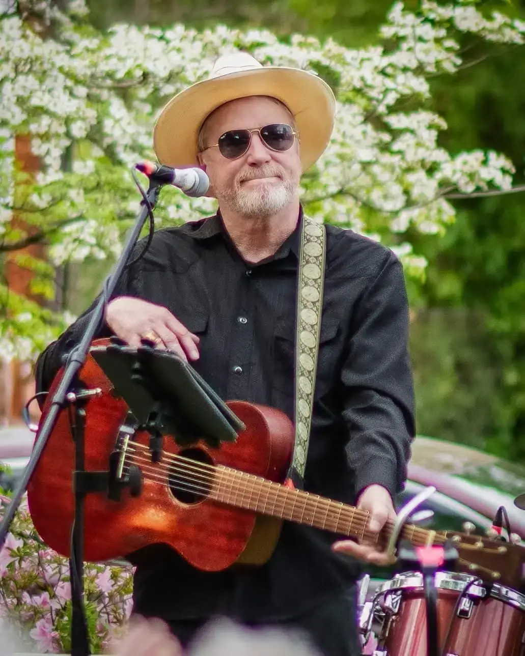 David Lowery wears a straw hat and sunglasses while playing an acoustic guitar during an outdoor performance.