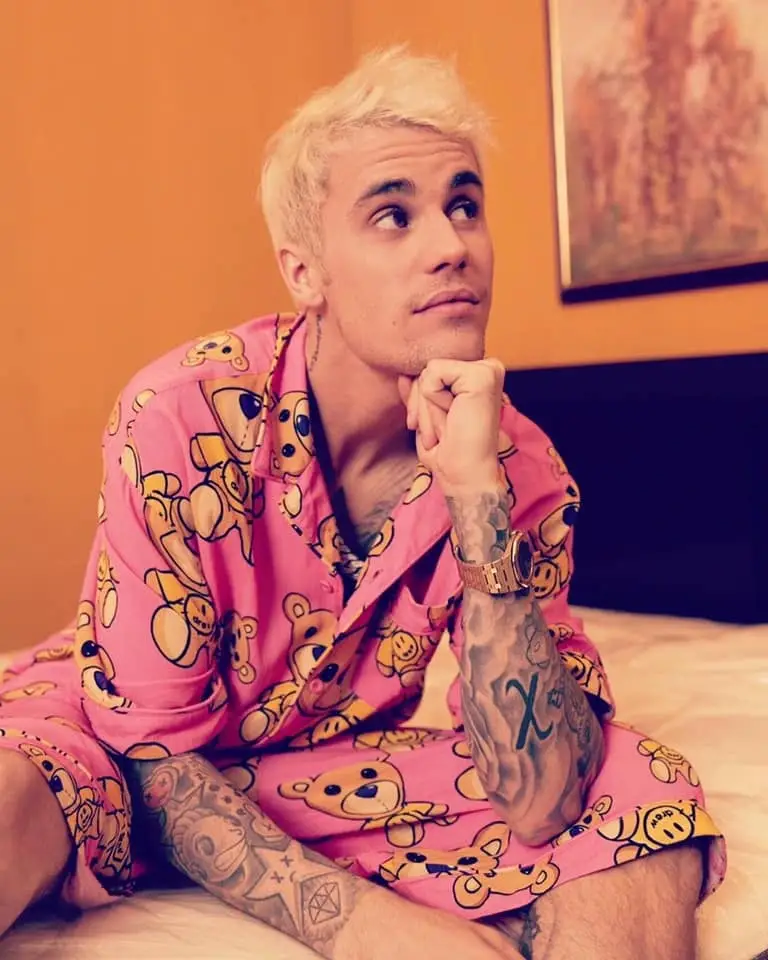 Justin Bieber sits on a bed wearing pink teddy bear print pajamas and a gold watch while looking upward.
