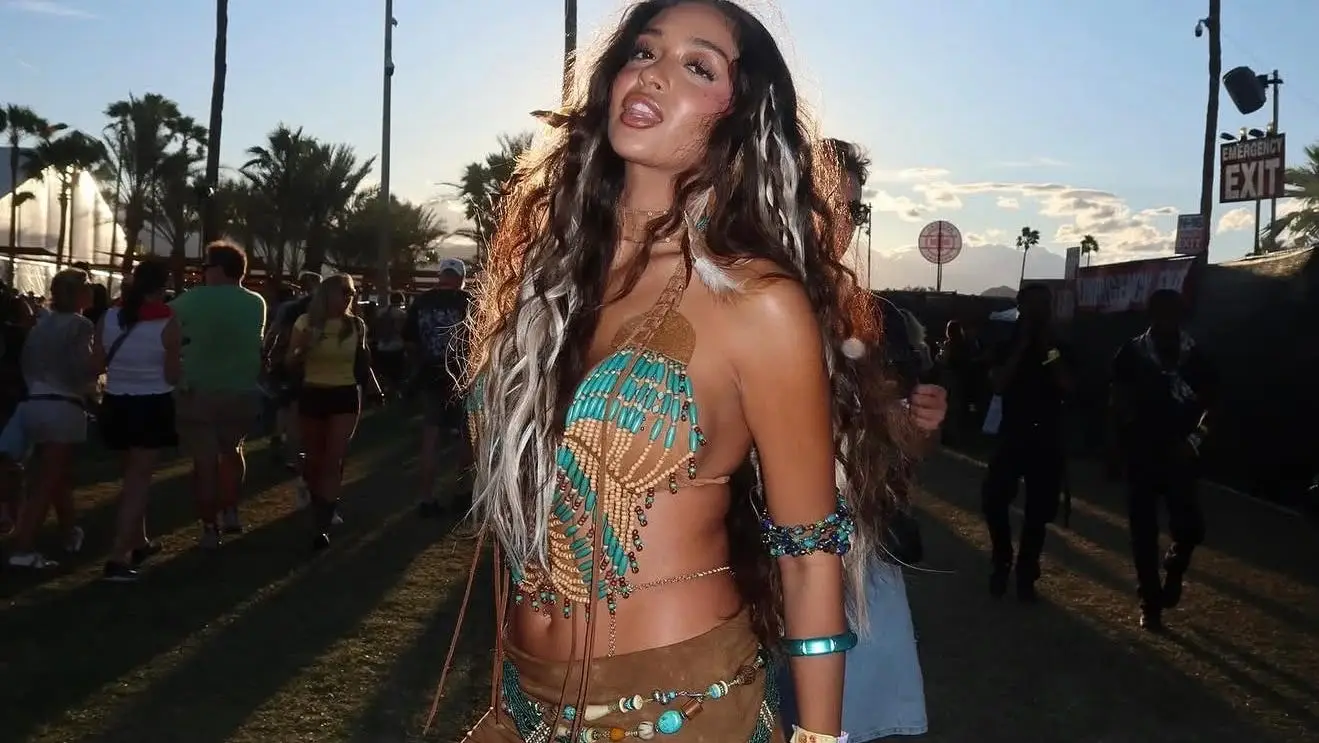 A woman poses at Coachella wearing a turquoise beaded fringe top and a bohemian-style brown skirt with feathered hair extensions.