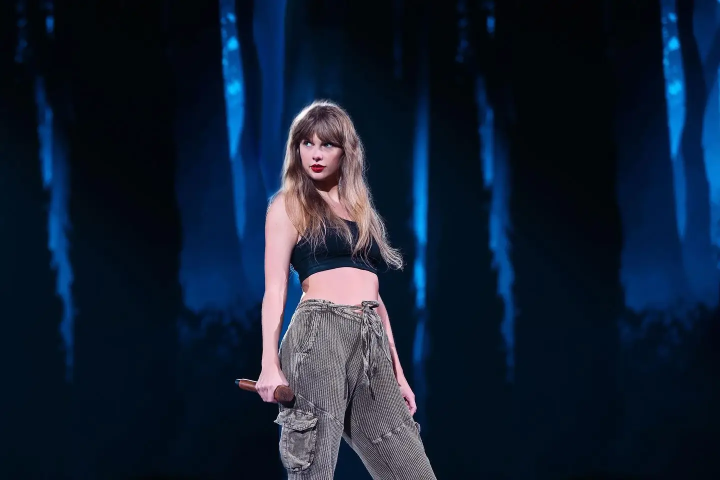 Taylor Swift performs on stage in a black crop top and olive cargo pants against a backdrop of glowing blue forest silhouettes.
