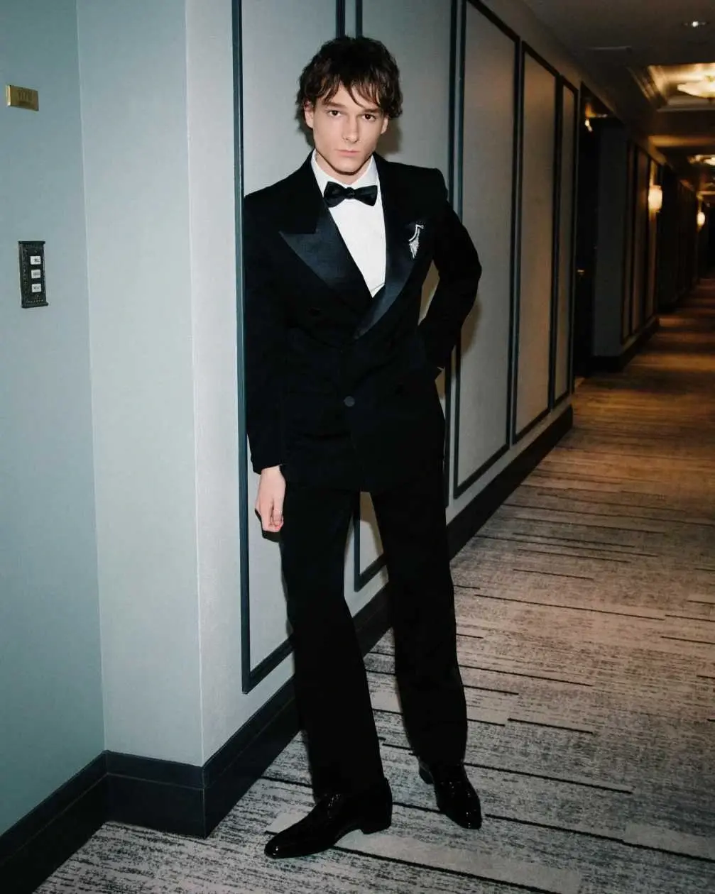 Mason Thames posing in a classic black tuxedo and bow tie while leaning against a hallway wall.