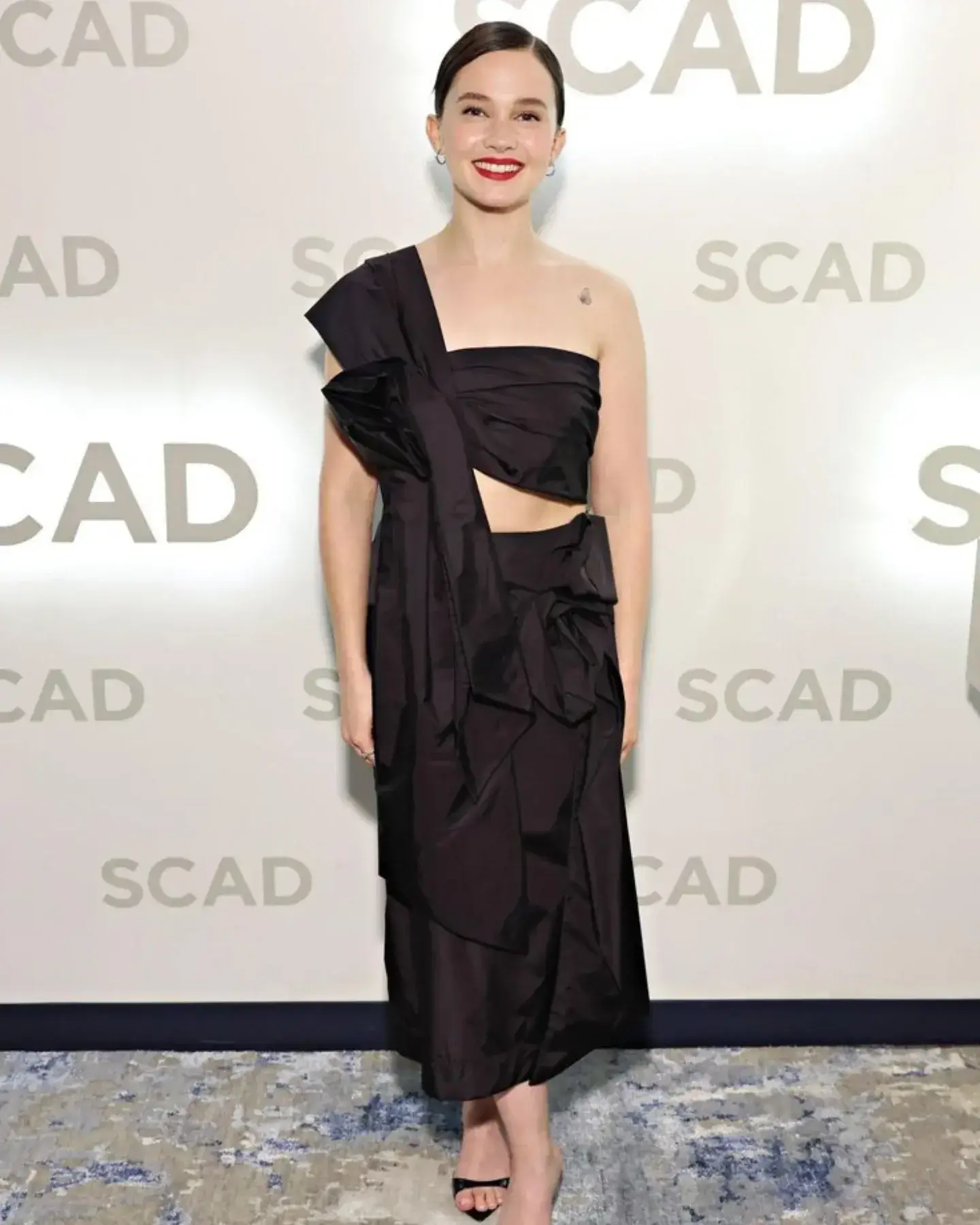 Cailee Spaeny poses on a red carpet wearing a structured black one-shoulder dress.