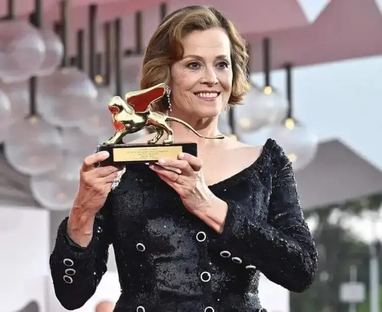 Sigourney Weaver smiling in a black sequined dress while holding the Golden Lion Honorary Award at the Venice Film Festival.