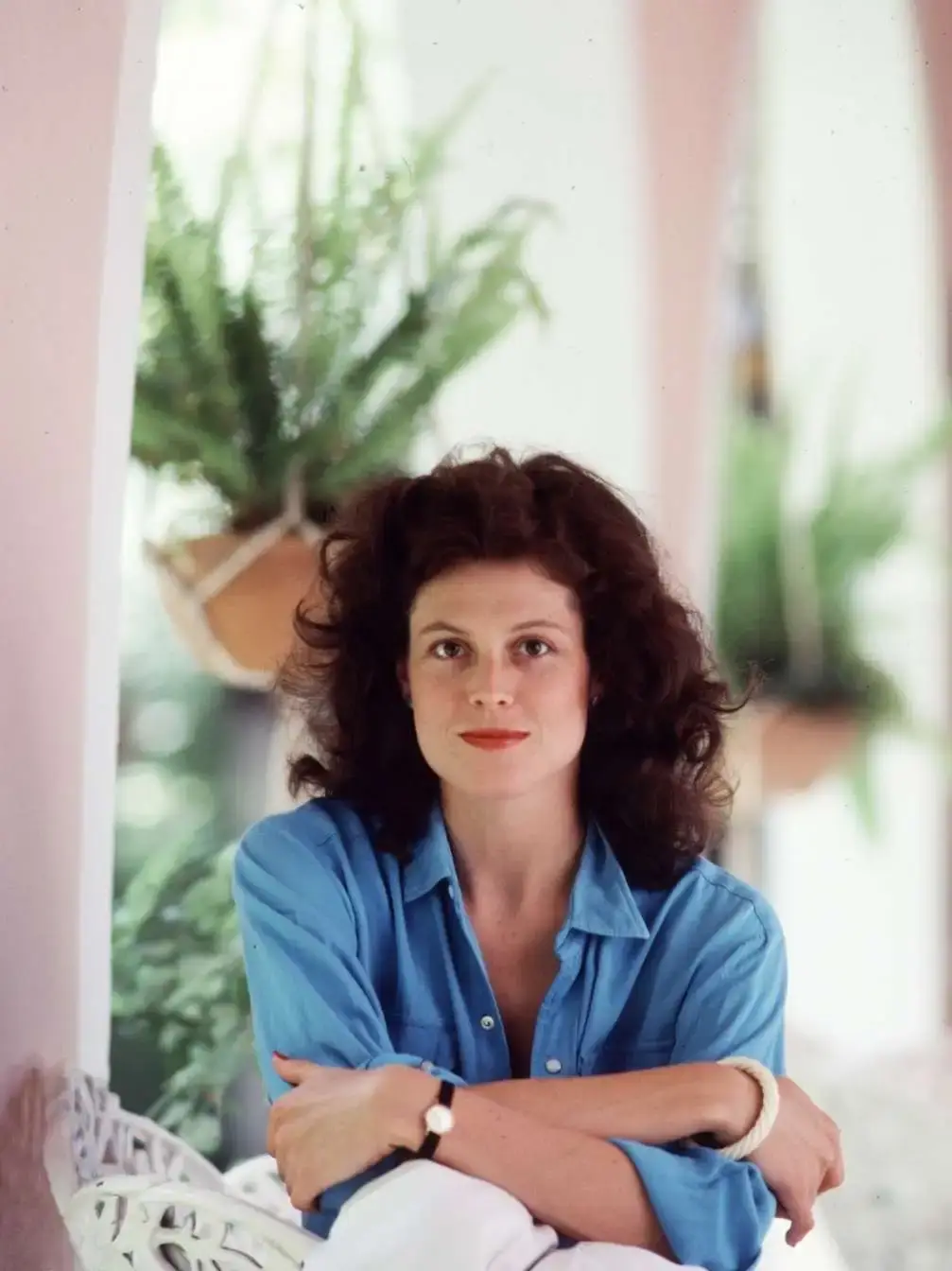 A vintage portrait of Sigourney Weaver with curly hair wearing a blue button-down shirt and sitting with her arms crossed.
